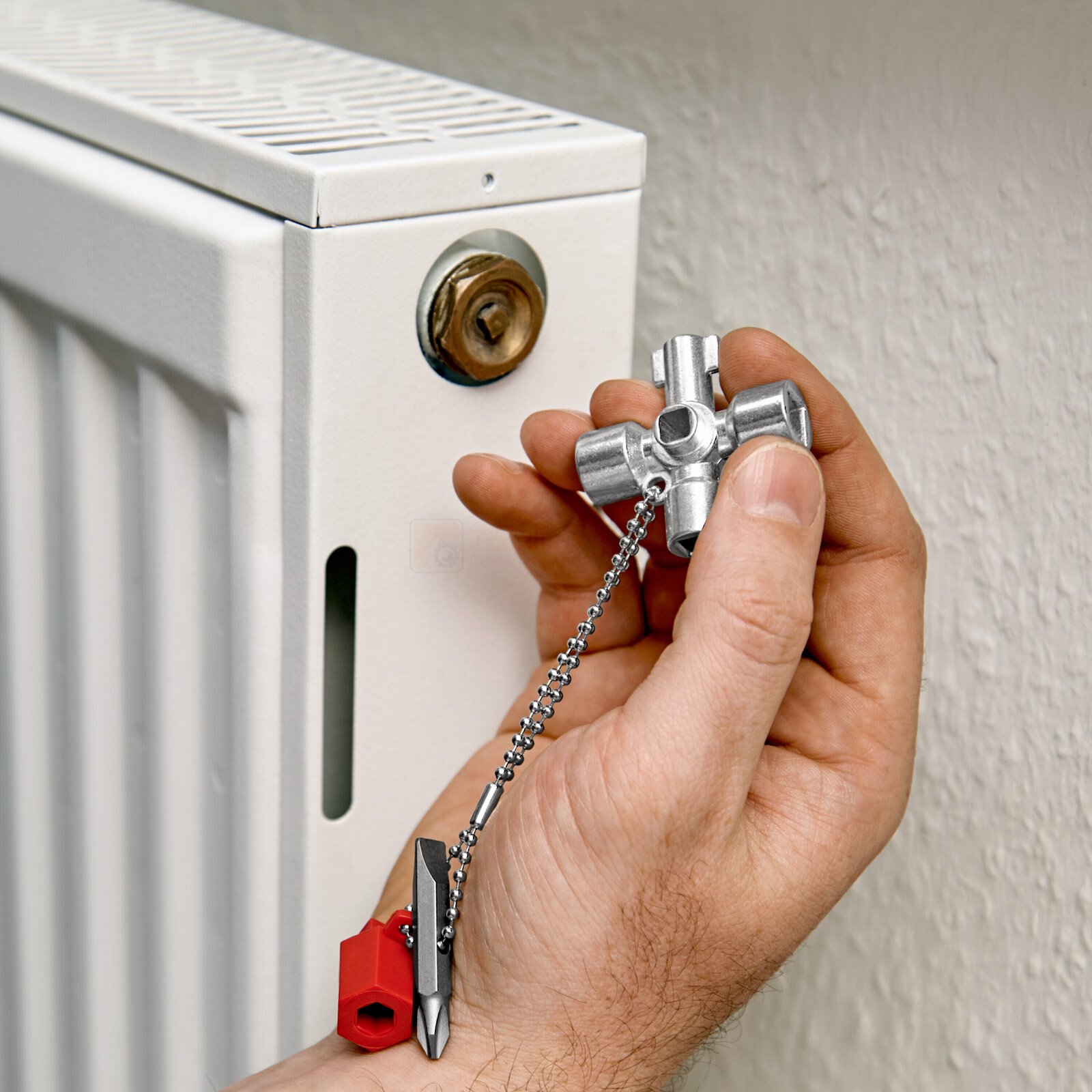 Pildil on käsi, mis hoiab Knipex standardvõtit hoonete tehniliste seadmete jaoks, millega on näha radiaatori õhutamist.