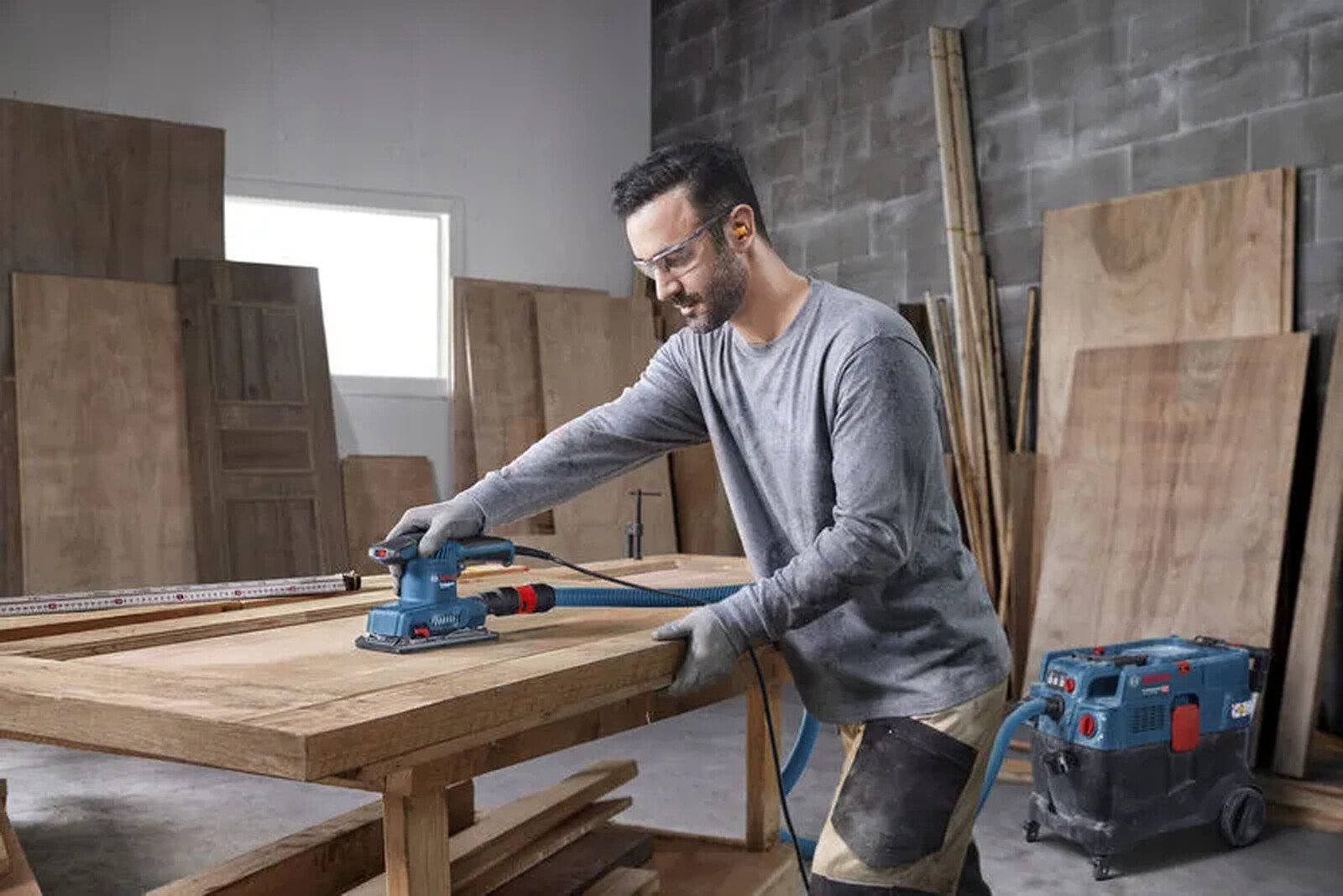 Pildil on mees, kes lihvib puidust pinda Bosch taldlihvija GSS 20-18 A-ga, kasutades tolmuimejat.