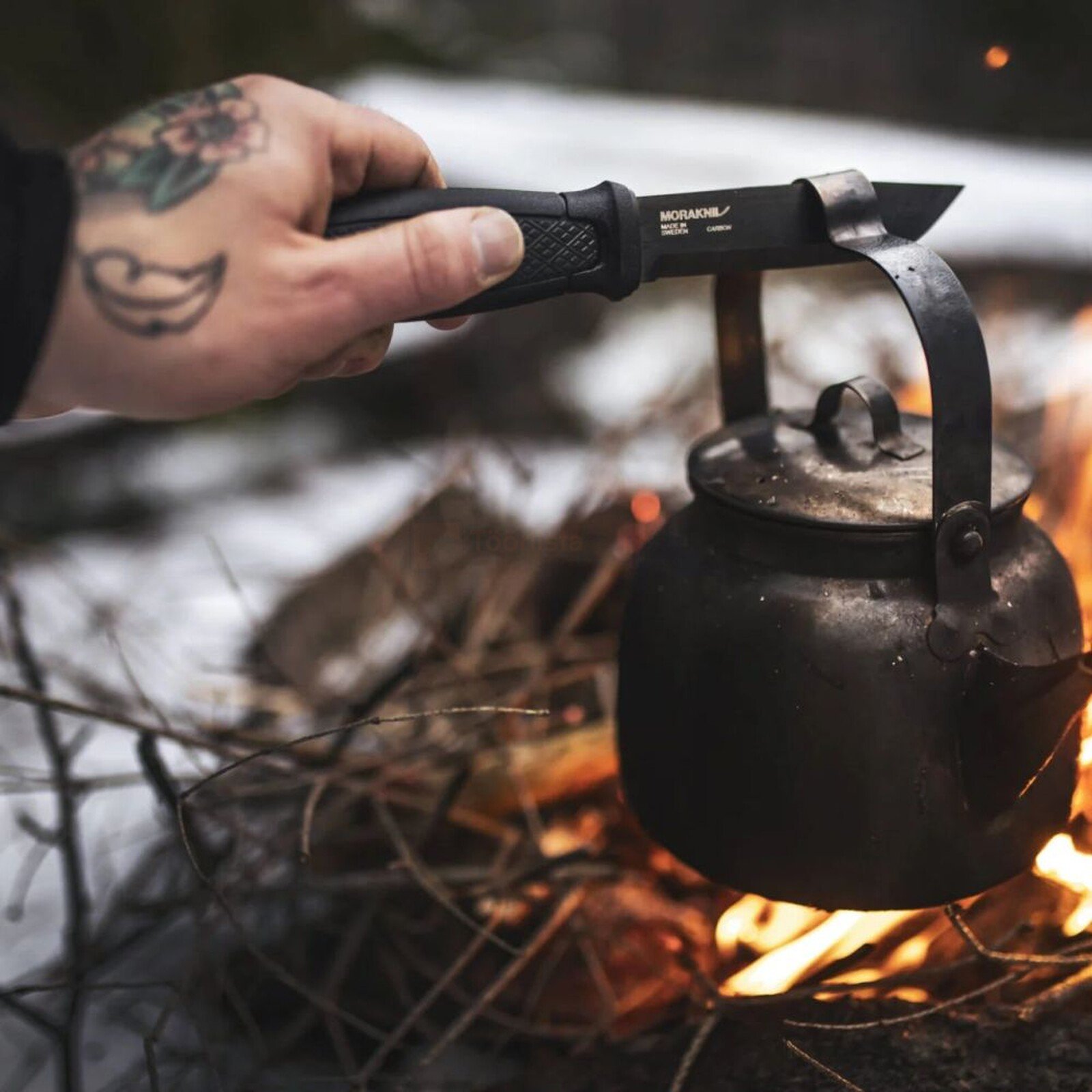 Pildil on käsi, mis hoiab MORAKNIV Garberg BlackBlade pussnuga, millega on riputatud vana veekeetja lõkke kohale.