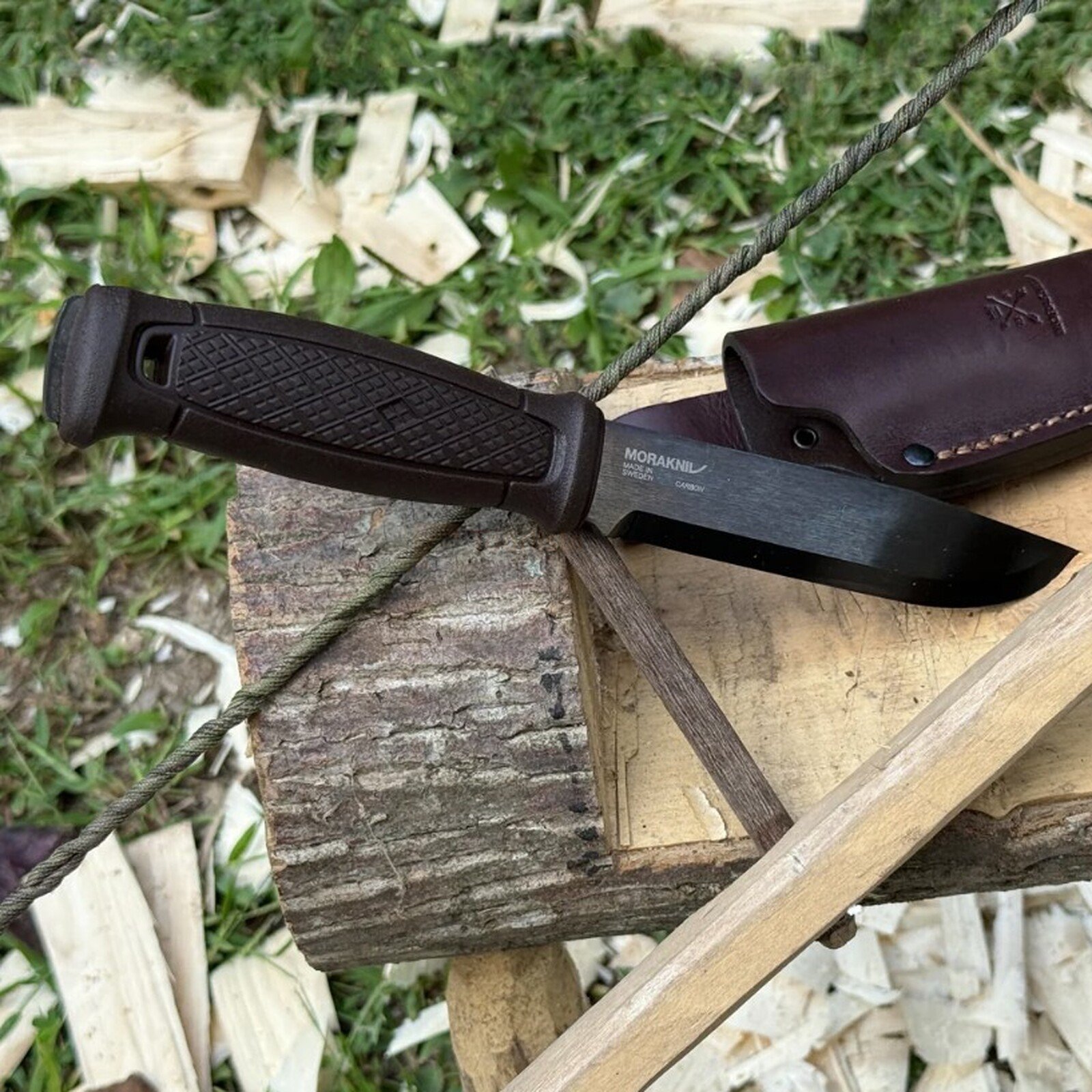 Pildil on näha musta teraga MORAKNIV Garberg Grand matkanuga koos pruuni nahktupaga, mis on asetatud puidust alusele...