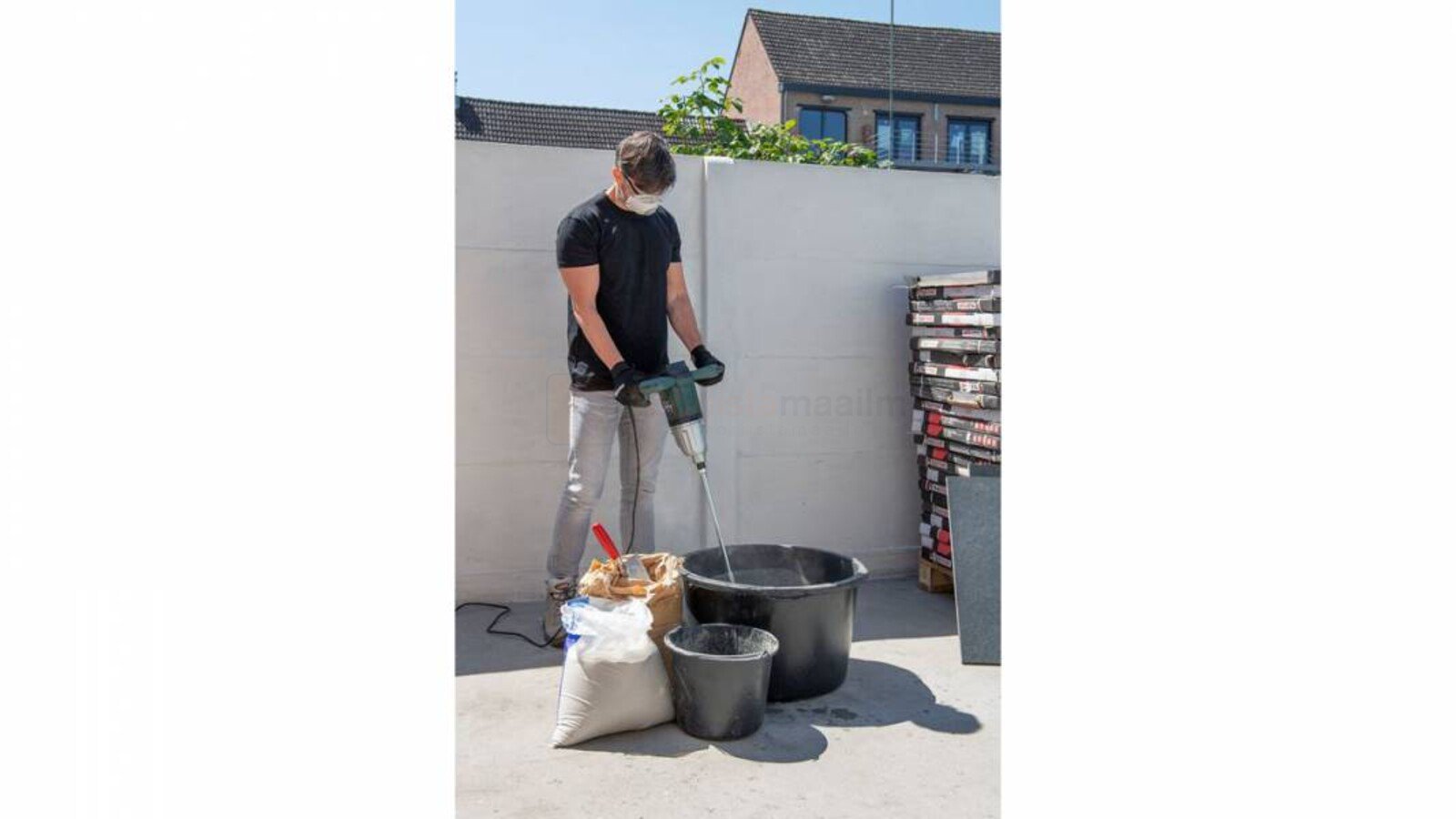 Pildil on mees, kes kasutab Powerplus E segumikserit POWP6040, et segada segu ämbris, mille kõrval on kott materjaliga.