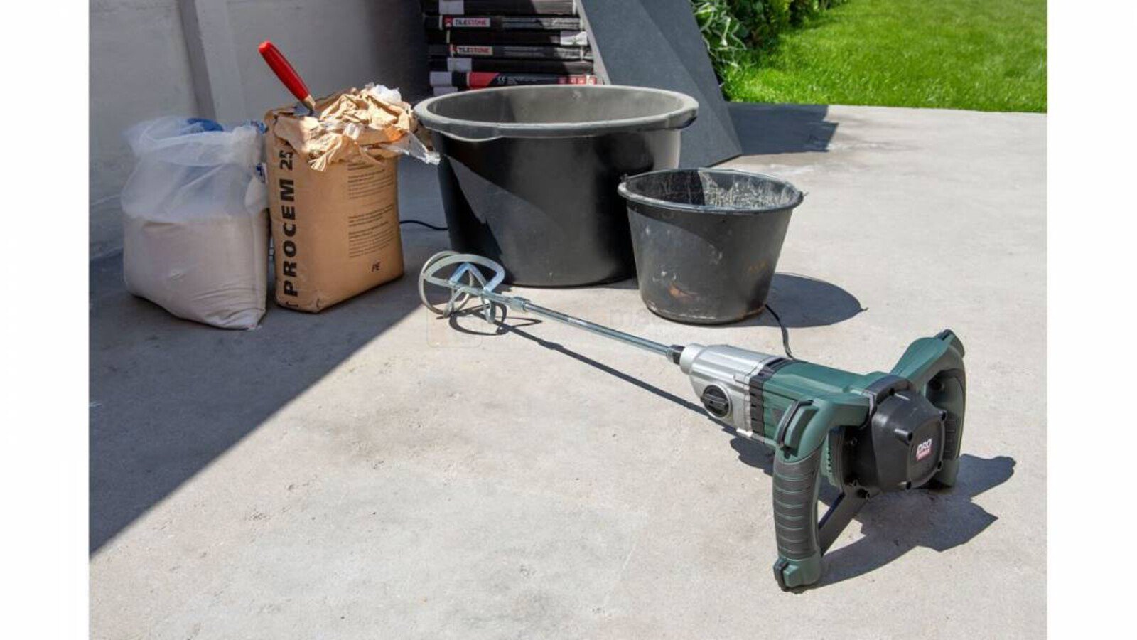 Pildil on segumikser Powerplus E POWP6040 koos segamismaterjalide, ämbrite ja labidaga betoonpõrandal.