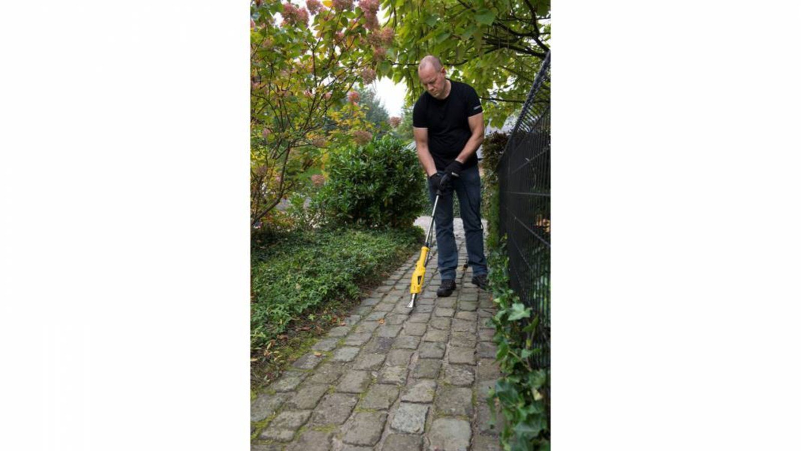 Pildil on mees, kes kasutab umbrohupõletit Powerplus X POWXG6611, et puhastada kivisillutist umbrohust.