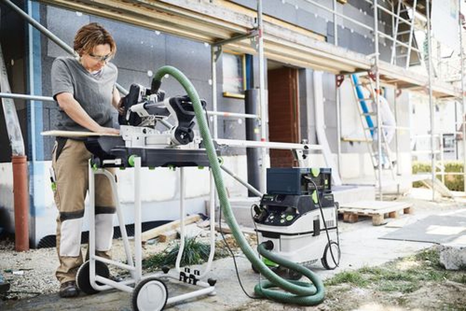 Pildil on ehitusplatsil töötav inimene, kes kasutab Festooli saagi koos tolmuimejaga, mis on ühendatud Festooli mobiilse...