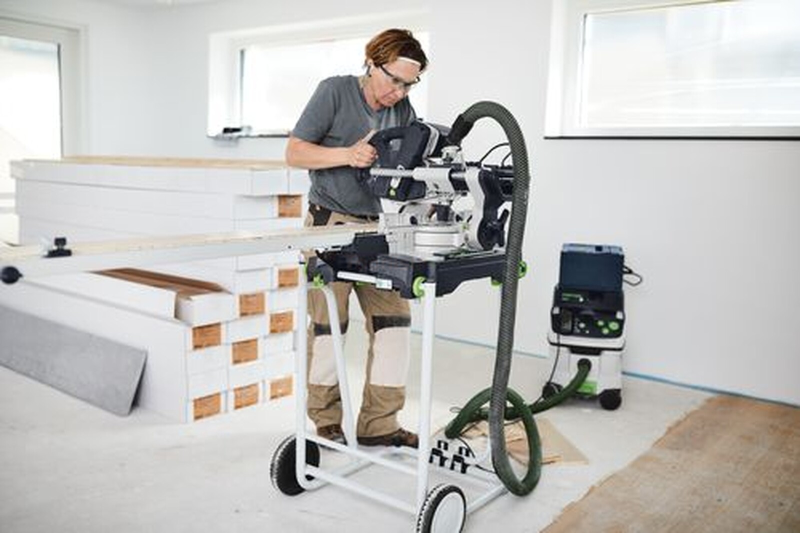 Pildil on mees, kes kasutab Festool saagi koos tolmuimejaga, mis on paigaldatud mobiilsele alusele, ning taustal on näha...