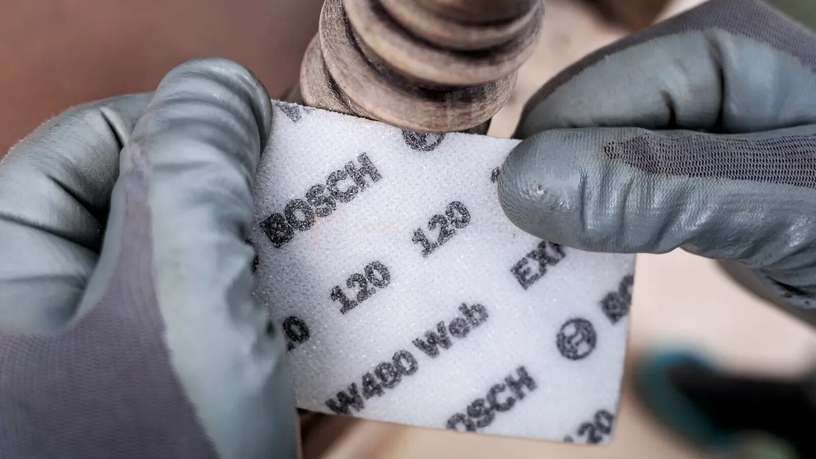 Pildil on näha, kuidas inimene hoiab kätega Bosch lihvvõrku käsilihvimiseks EXPERT W480 Web, et lihvida puidust detaili.