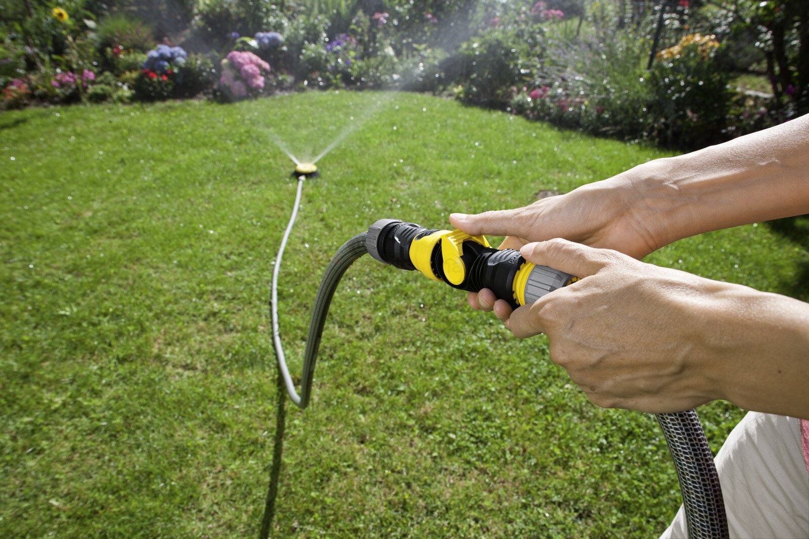 Pildil on näha, kuidas inimene hoiab käes Kärcheri juhtklappi, mis on ühendatud vooliku ja vihmutiga, et aeda kasta.