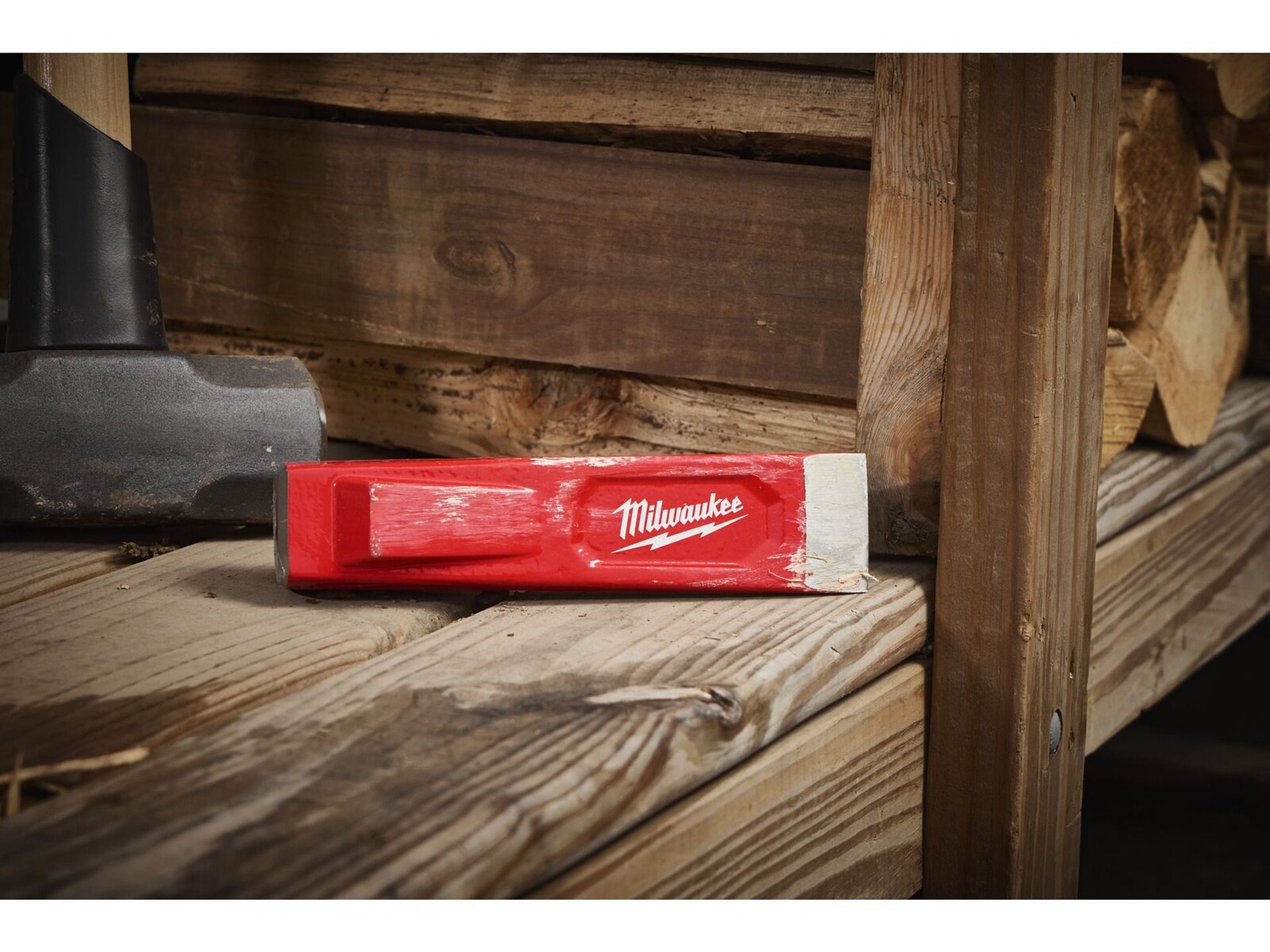 Pildil on Milwaukee punane 200 mm puulõhkumiskiil, mis on asetatud puidust alusele, mille taustal on näha puiduhunnik.