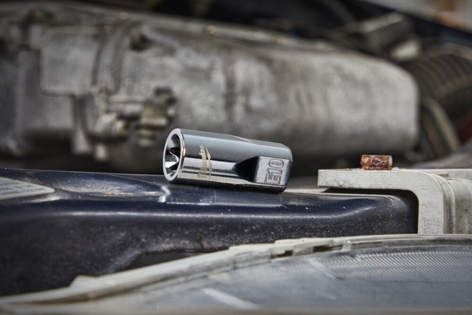 Pildil on Milwaukee E-Torx padrun, mis on asetatud auto mootoriruumi taustal.