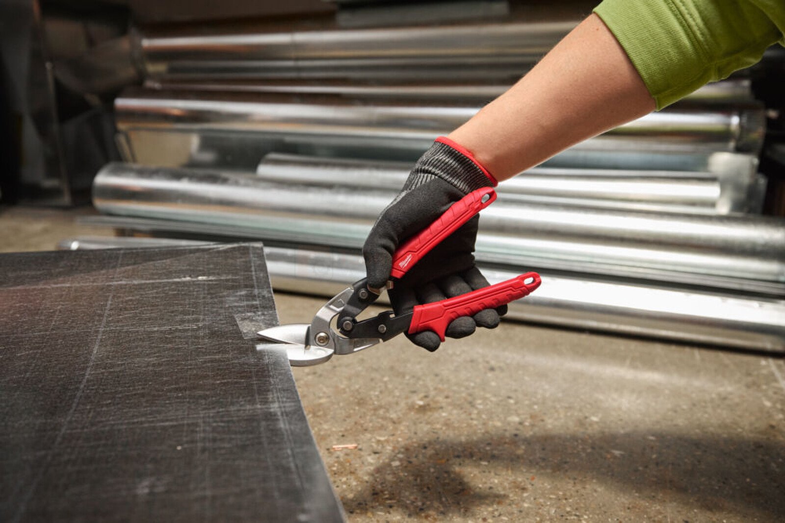 Pildil on käsi, mis kannab musti kindaid ja lõikab Milwaukee nihkega plekikääridega vasakule musta materjali.