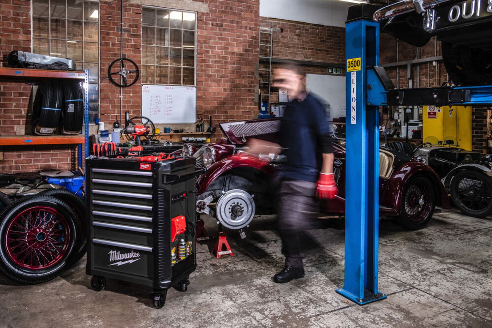 Pildil on Milwaukee tööriistakäru, mille kõrval on mehaanik, kes töötab punase klassikalise autoga garaažis.