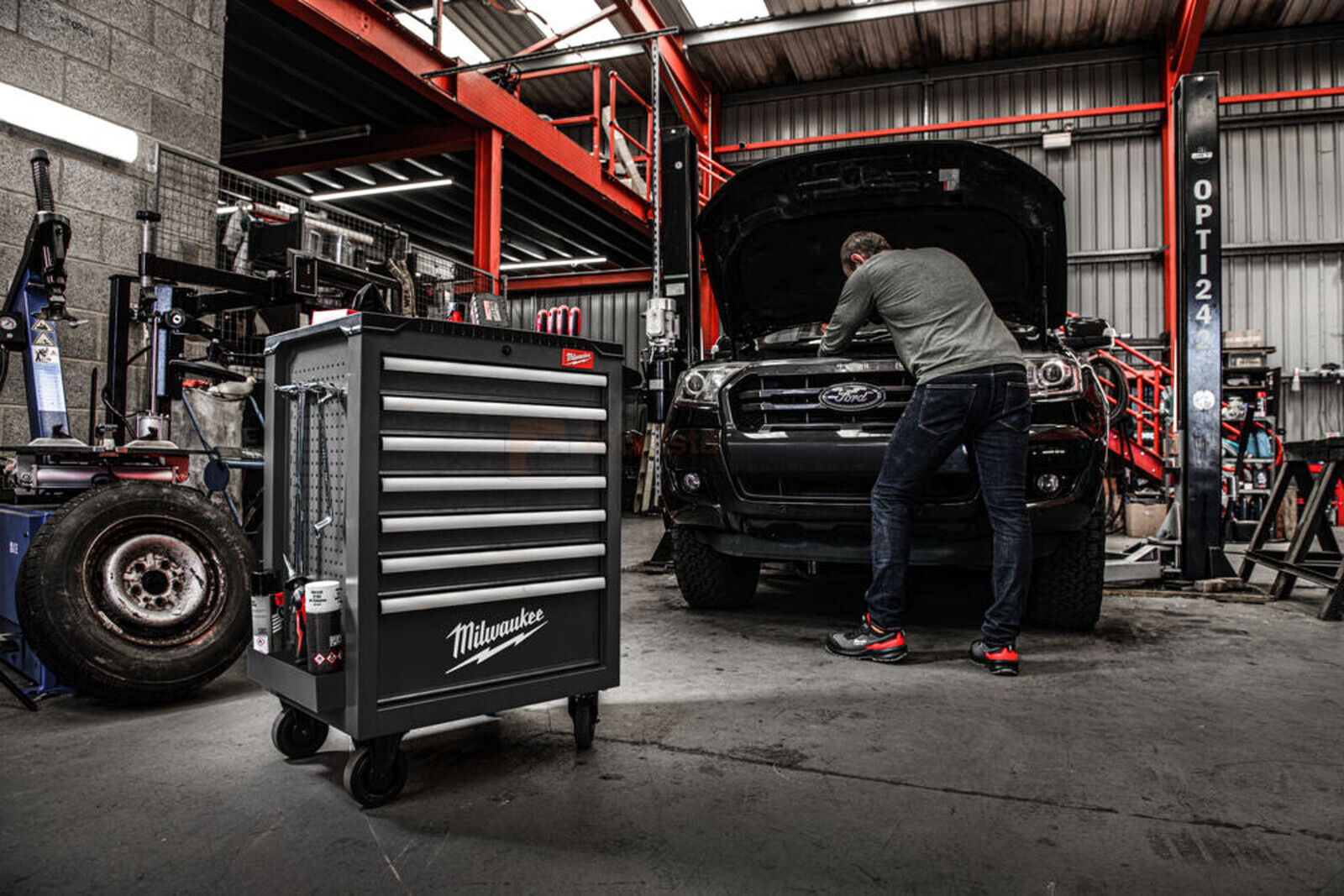 Pildil on Milwaukee tööriistakomplekt halli tööriistakäruga, mille kõrval töötab mehaanik auto kallal töökojas.