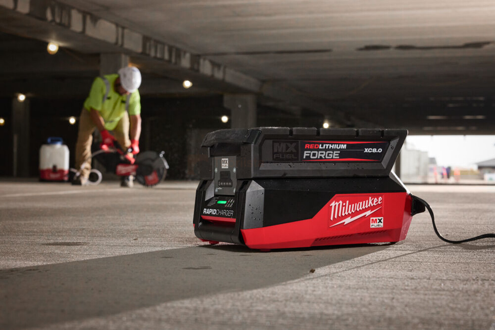 Pildil on Milwaukee kiirlaadija MXF FC, mis asub betoonpõrandal ja mille taustal töötab ehitustööline.