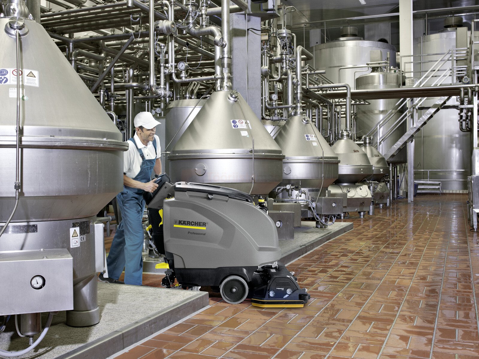 Pildil on töötaja, kes kasutab Kärcheri põrandapuhastusmasinat tehase keskkonnas, kus on näha suuri mahuteid ja torustikku.
