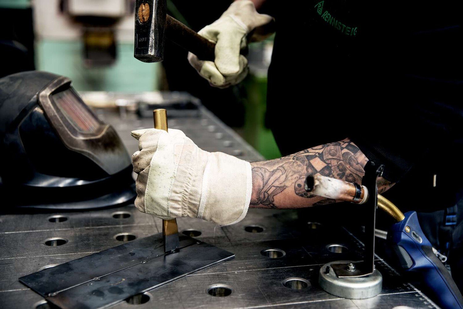Pildil on mees, kes kasutab Rennsteig lamemeislit metalli töötlemiseks, samal ajal kui tema kõrval on keevitusmask.