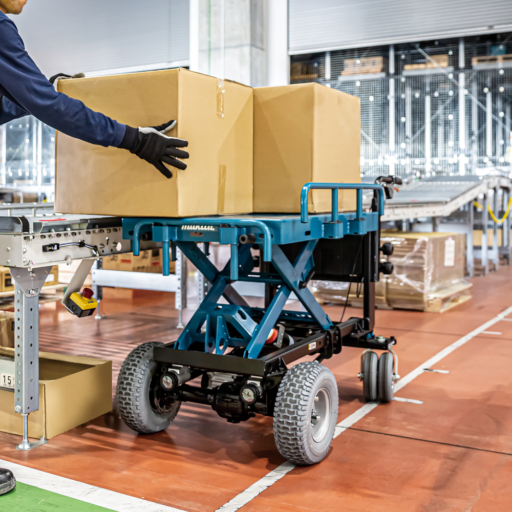 Pildil on Makita akutoitega tõstuk-käru DCU601Z, ilma aku ja laadijata, millega töötaja asetab karbid platvormile.