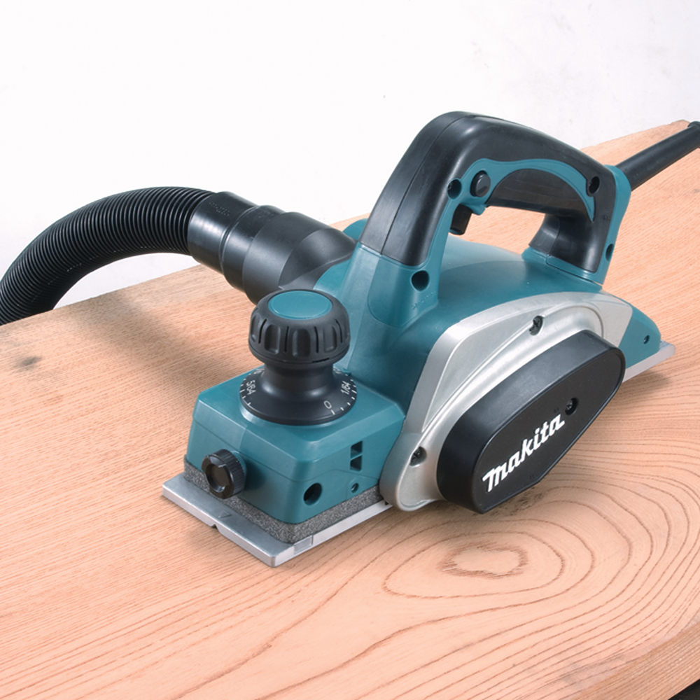 Pildil on Makita KP0800J höövel, mis on asetatud puidule ja ühendatud tolmuimeja voolikuga.