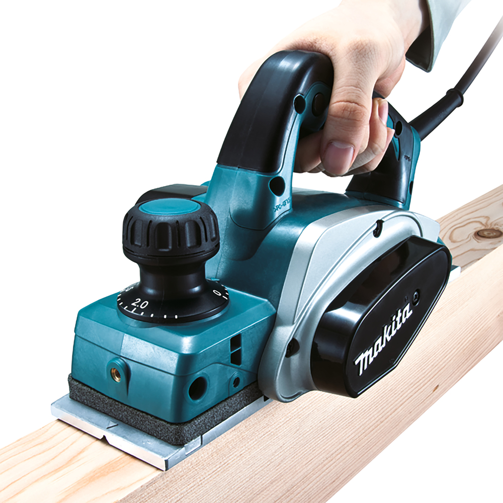 Pildil on näha Makita KP0800J höövel, mida kasutaja hoiab ja kasutab puidu hööveldamiseks.