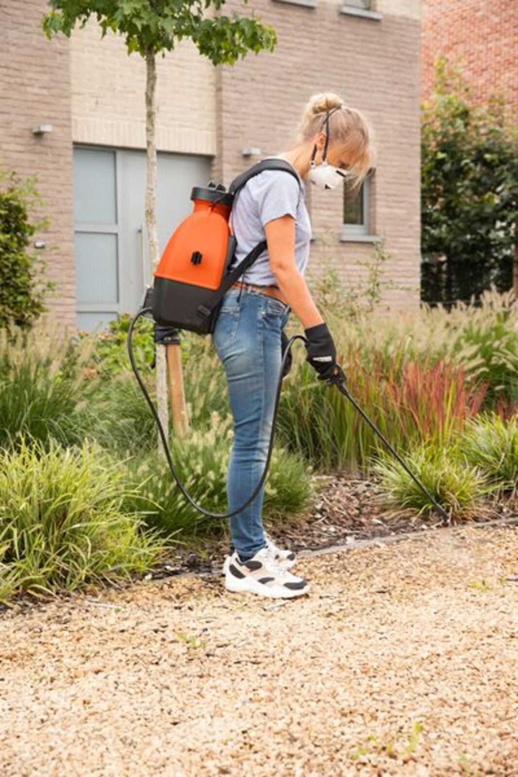 Pildil on naine, kes kasutab Powerplus Dualpower akutoitel survepihustit, kandes kaitsemaski ja kindaid, et pritsida...