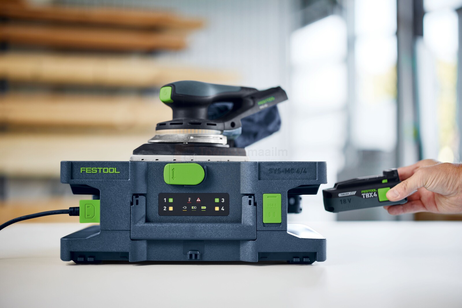 Pildil on Festool kiirlaadija SYS-MC 6/4, mille peal on kinnitatud lihvija ja mille kõrval on käsi, mis sisestab akut.