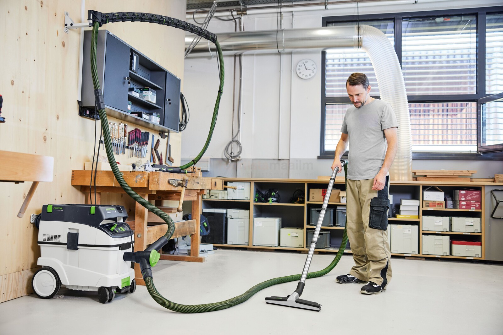 Pildil on mees, kes kasutab Festooli tolmuimejat koos Y-ventiiliga, et puhastada töökoja põrandat.