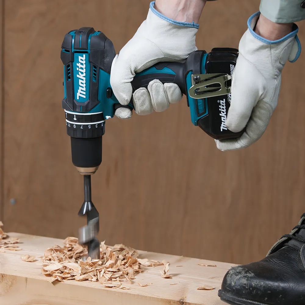 Pildil on Makita akulööktrell DHP482Z, mis puurib puitu, koos kasutaja kätega kinnastega ja jalanõuga.