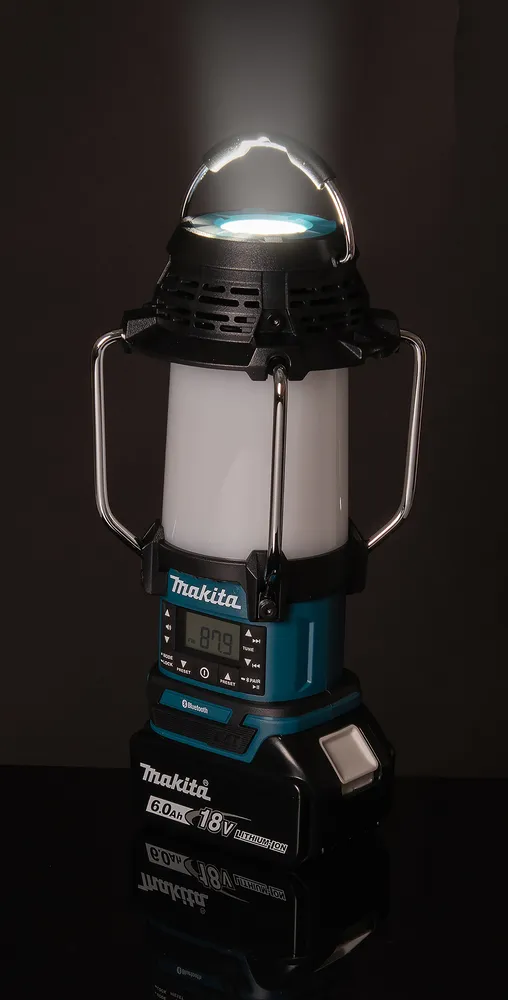 Pildil on Makita DMR057 akulamp-raadio, millelt paistab eredalt valgus, ja see on asetatud läikivale pinnale, peegeldudes...