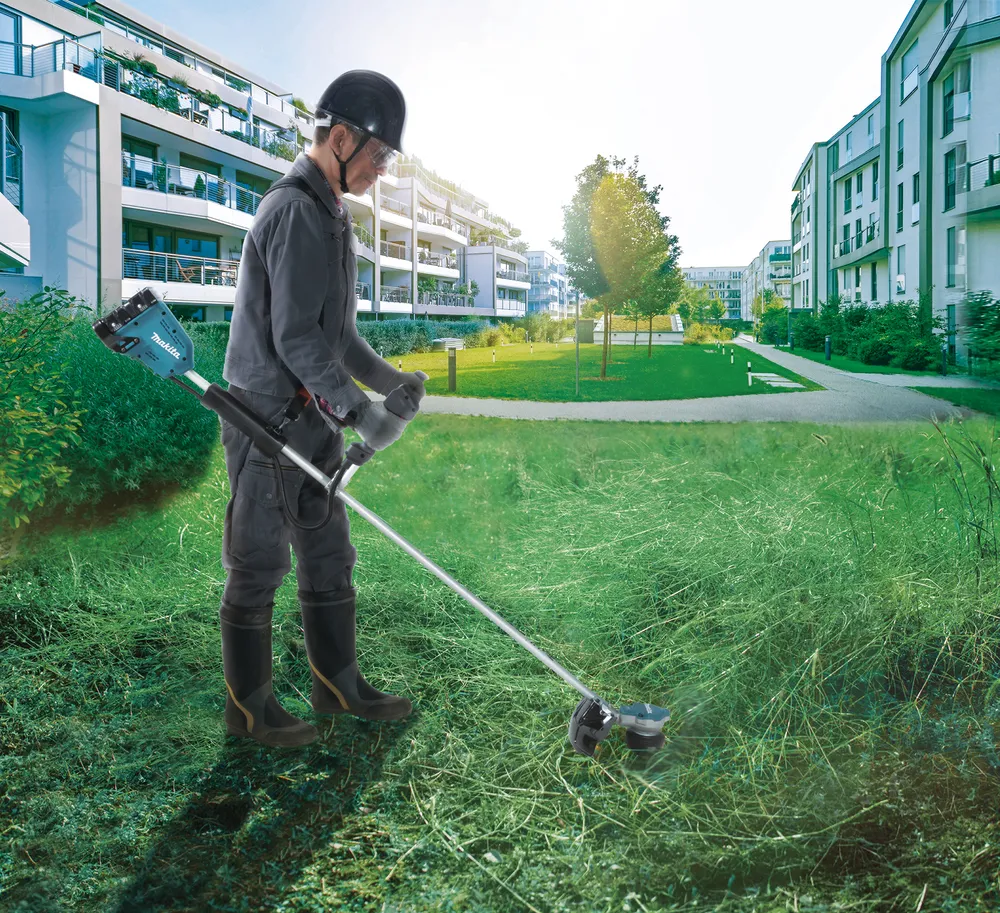 Pildil on Makita akutrimmer DUR368LZ, mida inimene kasutab muru niitmiseks uusehitiste vahelises hooldatud keskkonnas.