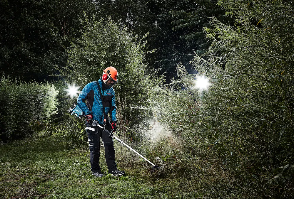 Pildil on Makita vahetatavate tarvikutega akutrimmer DUX60Z, mida mees kasutab õhtuhämaruses heki kärpimiseks.