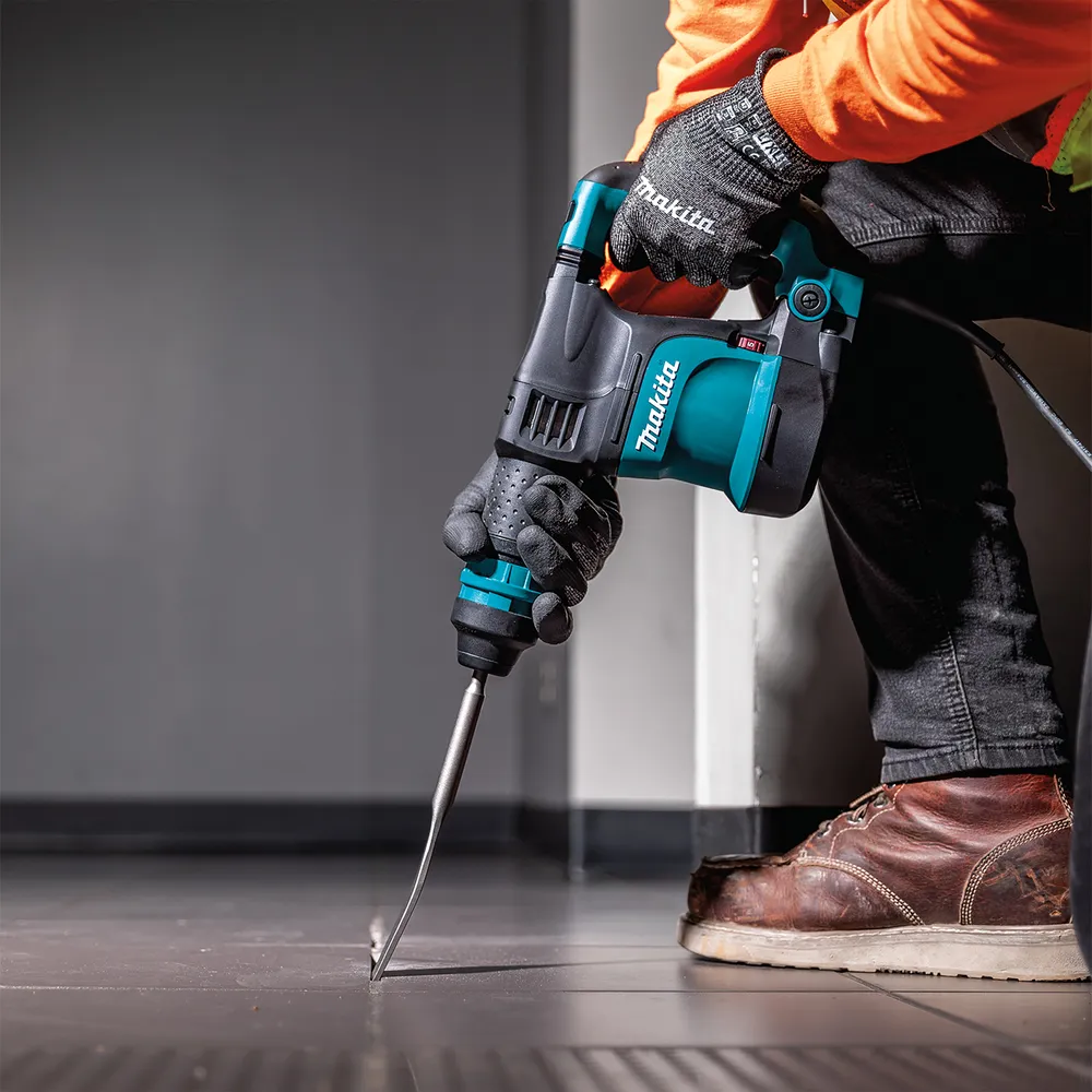 Pildil on Makita piikvasar HK1820 SDS-Plus, mida inimene hoiab tööriistakindaga käes ja kasutab seda õues töö tegemiseks.