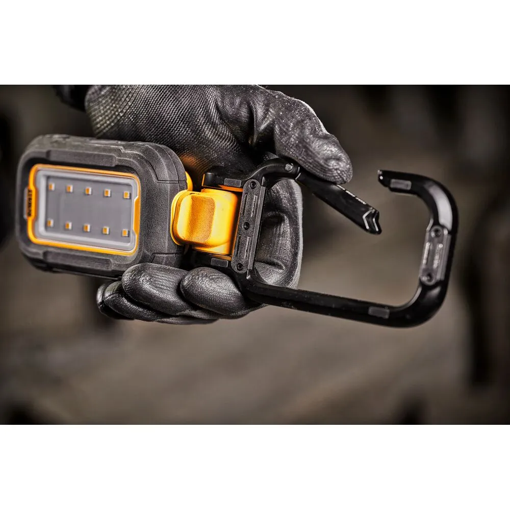 Pildil on DeWalt laetav USB-C LED töövalgusti, millel onintegreeritud karabiinkonks, mida hoitakse töökindaid käes.